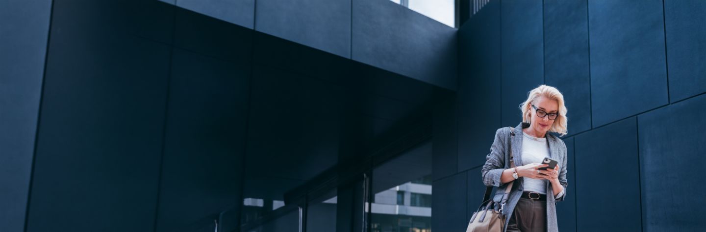 A blonde businesswoman in a grey blazer is standing outside a modern building, looking at her smartphone and smiling.
