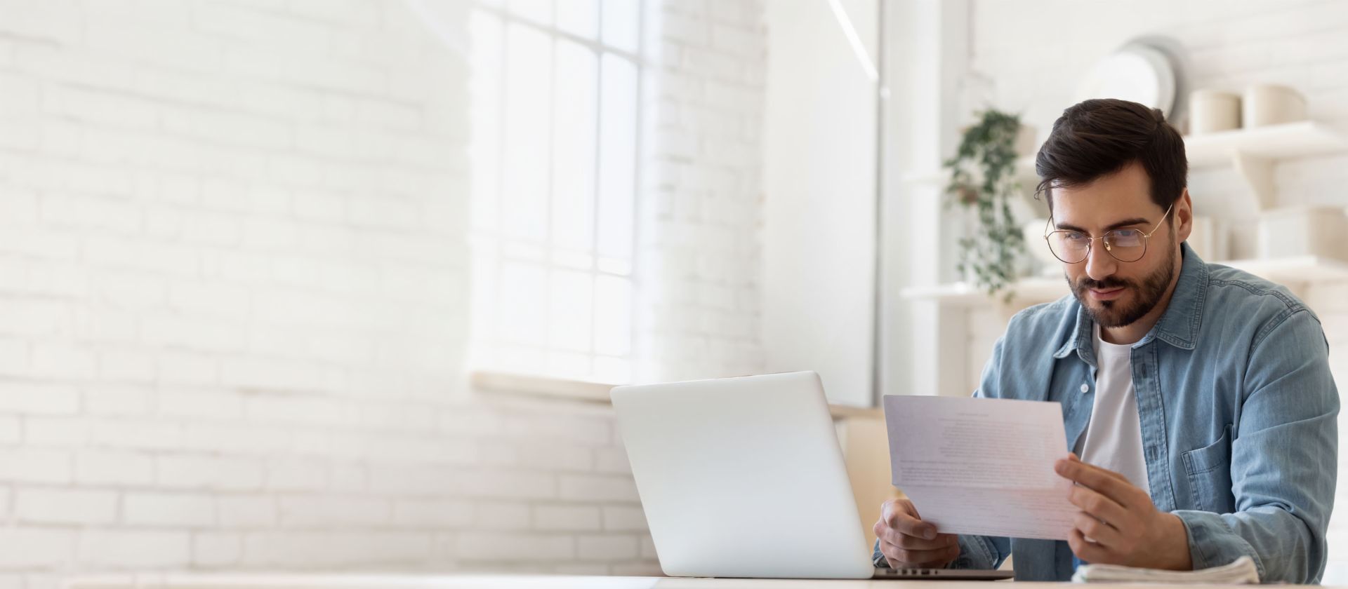 Man wearing glasses working on laptop while reviewing debt settlement document in bright home 