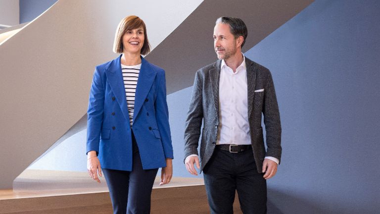 The CEO in a gray blazer and the CFO of the EOS Group in a blue blazer walk through a modern office. The space features large windows, a curved wall, and stylish hanging lamps.