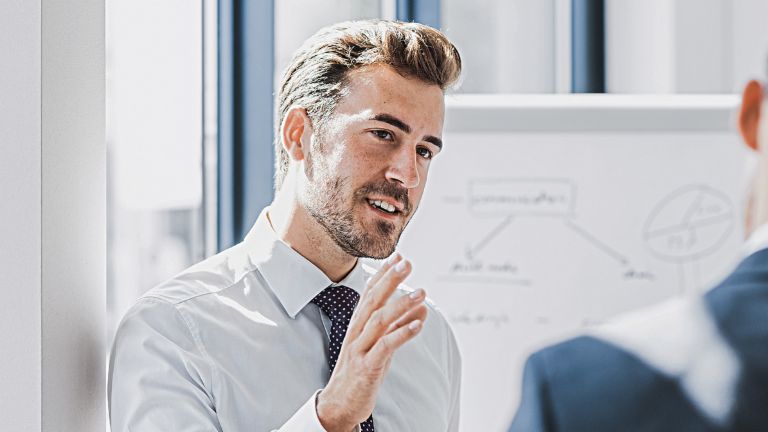 You can see a man in business attire explaining something with a friendly, engaged expression. In front of him stands another person who is clearly listening to him. You can see a whiteboard with a sketch in the background.