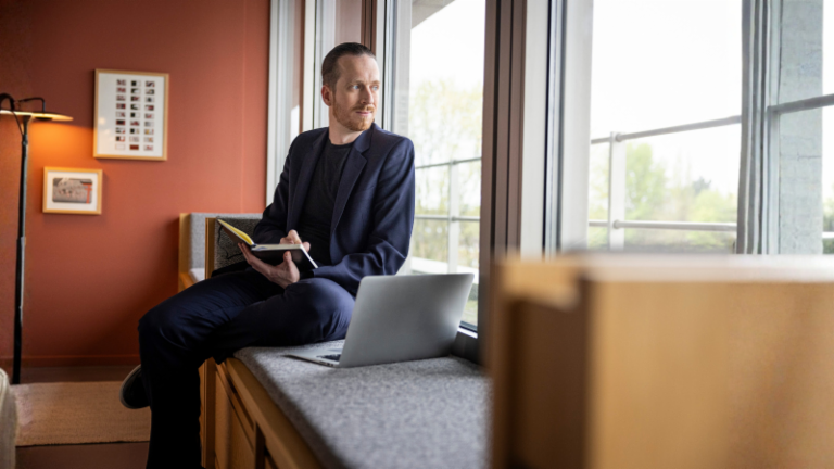 Sebastian Pollmer sits on a windowsill with an open laptop and a notebook in his hand, staring out the window.