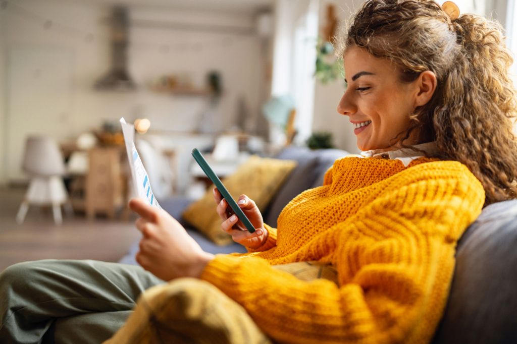 A smiling young woman with curly hair, wearing a cozy yellow sweater, sits on a couch in her living room, looking at her smartphone while holding a paper document.