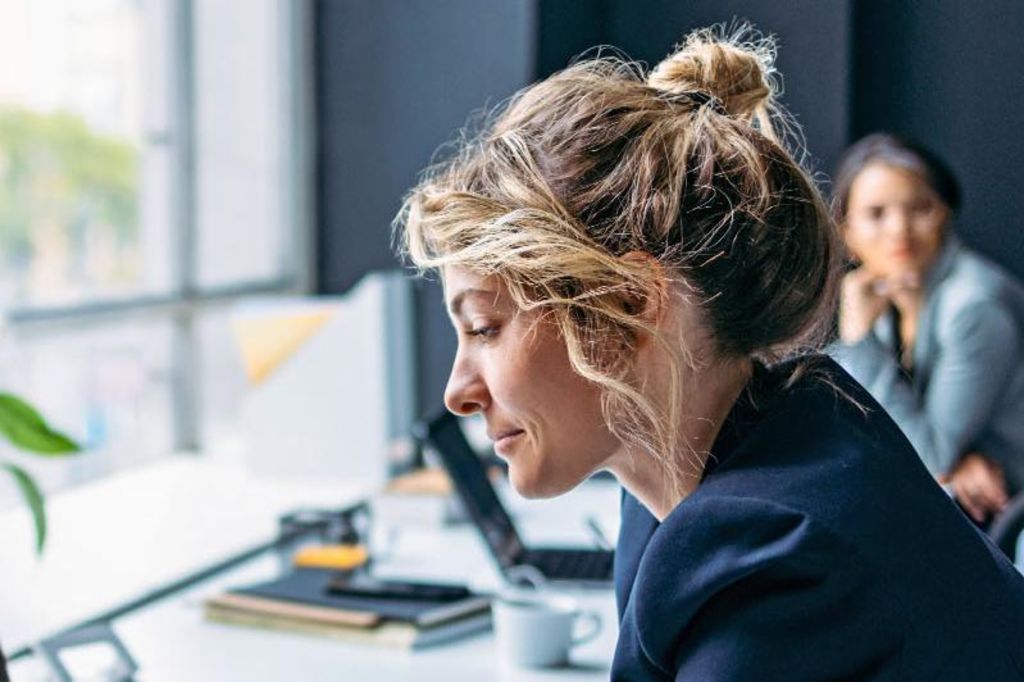 The image shows a woman sitting at a desk in a modern office, wearing a dark blazer while focusing on her laptop, with a plant and some office supplies visible on the table, and another person sitting in the background.