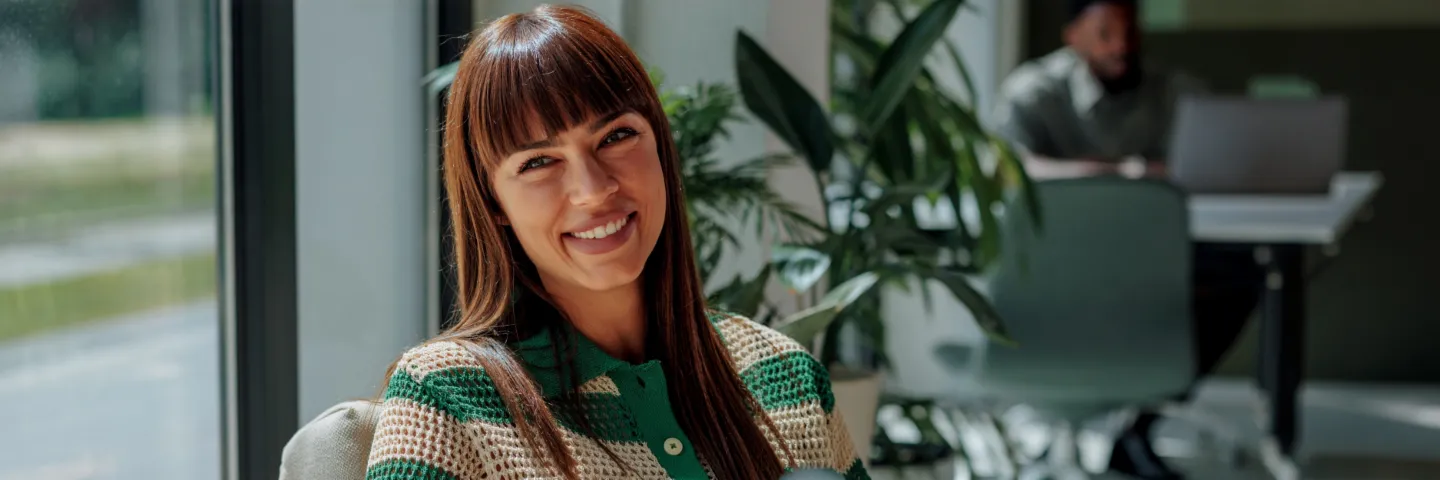 A smiling woman with dark hair sits comfortably in a bright, modern office and looks warmly into the camera. In the background, other people and large windows are visible.