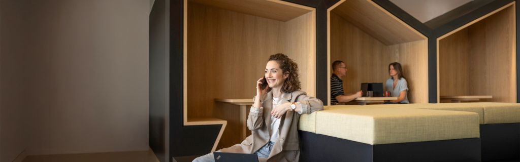 A smiling person sits in a modern office lounge, talking on the phone and working on a laptop. In the background, two other people are working.