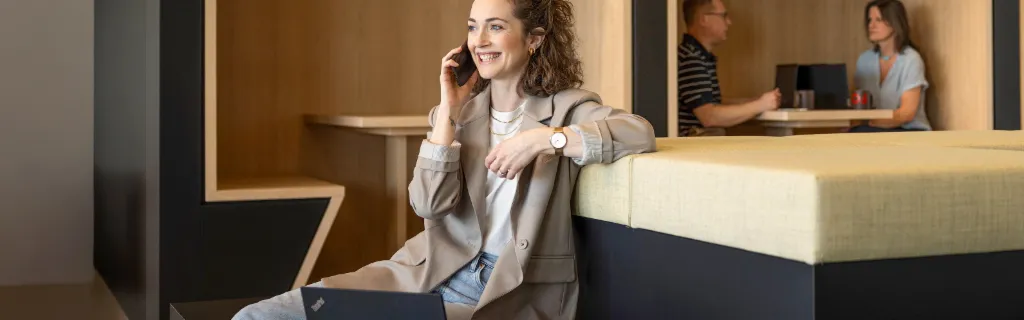 A smiling person sits in a modern office lounge, talking on the phone and working on a laptop. In the background, two other people are working.