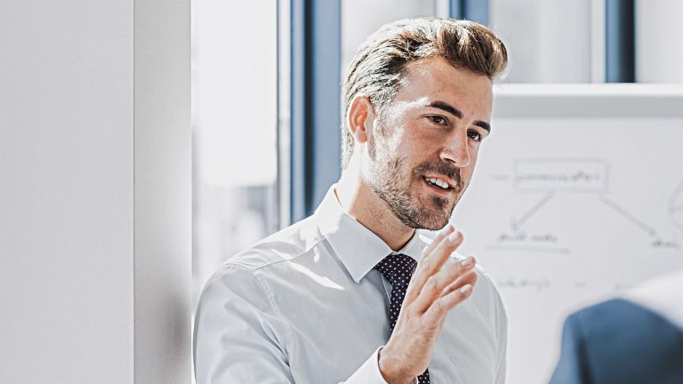 You can see a man in business attire explaining something with a friendly, engaged expression. In front of him stands another person who is clearly listening to him. You can see a whiteboard with a sketch in the background.