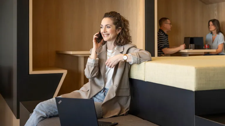 A smiling person sits in a modern office lounge, talking on the phone and working on a laptop. In the background, two other people are working.