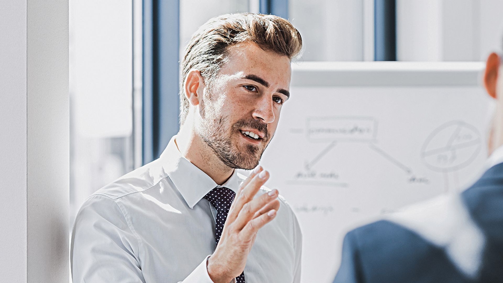 You can see a man in business attire explaining something with a friendly, engaged expression. In front of him stands another person who is clearly listening to him. You can see a whiteboard with a sketch in the background.