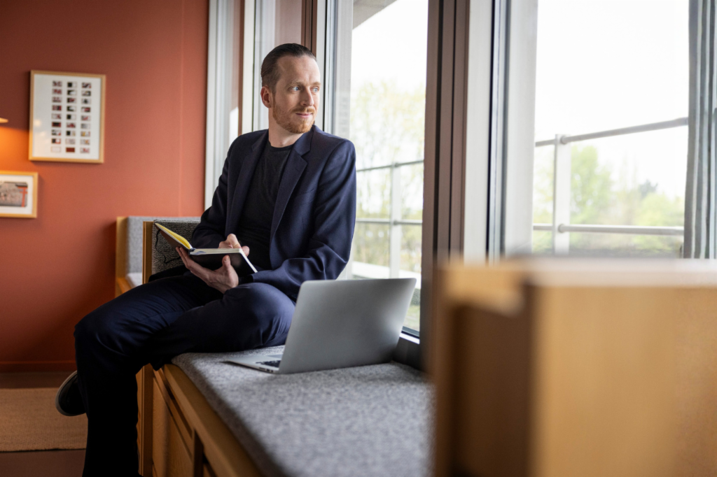 Sebastian Pollmer sits on a windowsill with an open laptop and a notebook in his hand, staring out the window.