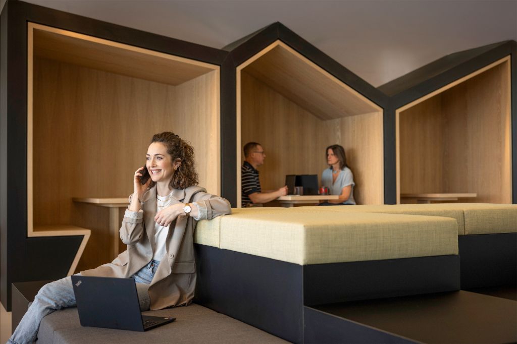 A smiling person sits in a modern office lounge, talking on the phone and working on a laptop. In the background, two other people are working.