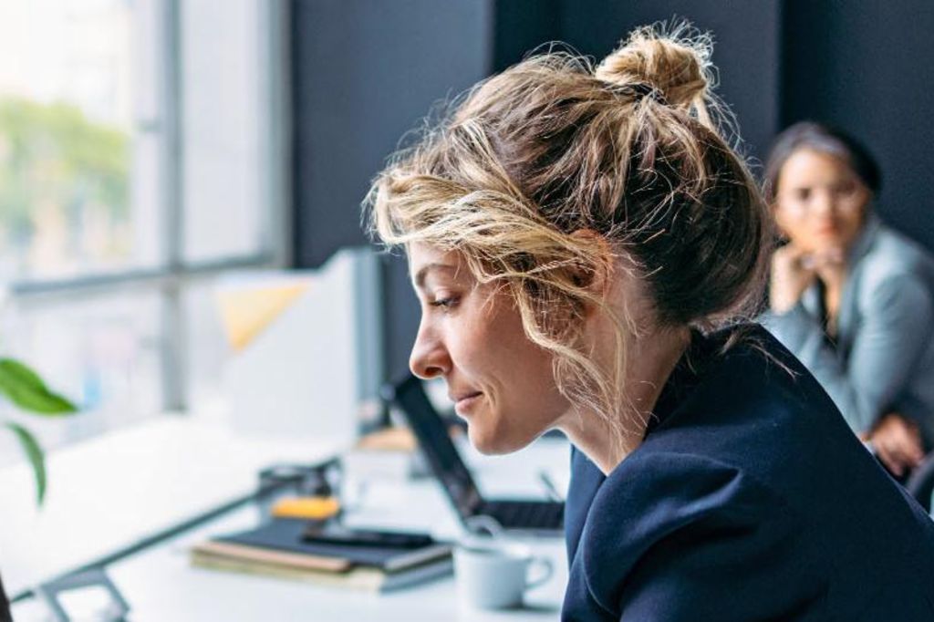 The image shows a woman sitting at a desk in a modern office, wearing a dark blazer while focusing on her laptop, with a plant and some office supplies visible on the table, and another person sitting in the background.