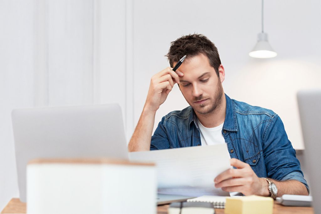 Man sitting at the living room table looking seriously at bills lying next to his laptop. 